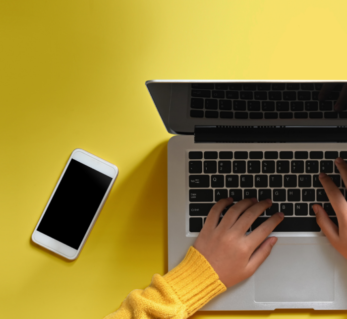 Photo of a laptop and mobile phone with a yellow background and some hands over the keyboard looking like the person is typing. 