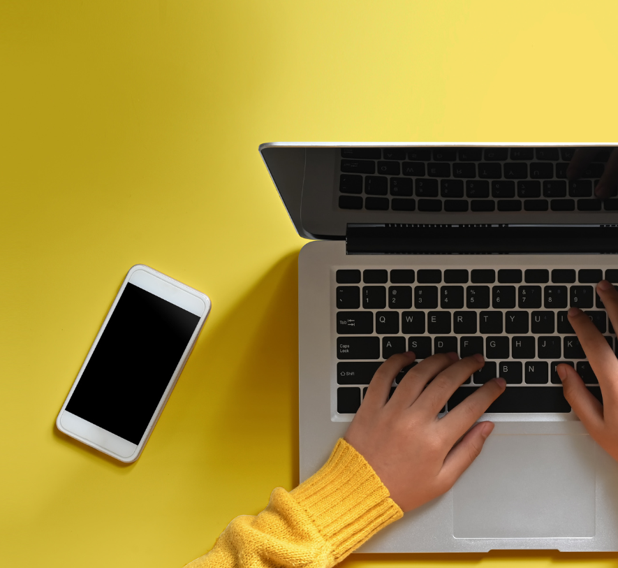 Photo of a hand typing on a laptop, a mobile phone is placed next to the laptop and there is a yellow background. Photo is for decorative purposes only. 