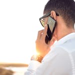 Man outdoors in Morocco talking on smartphone, wearing sunglasses and white shirt, backlit by sunset.